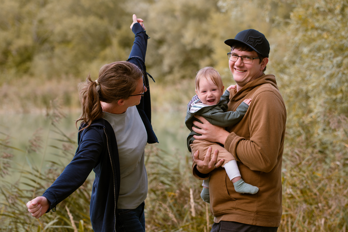 Mama streckt fröhlich die Arme in die Höhe, während Papa ihr lachendes Baby im Arm hält und beide Richtung Kamera lächeln.
